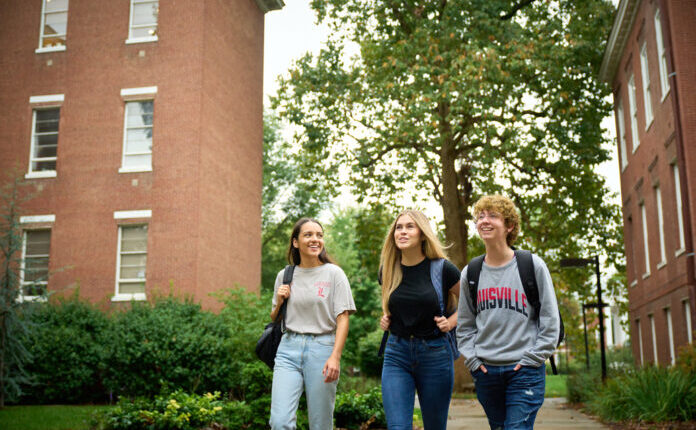 Three students wearing UofL shirts and carrying backpacks walk on a sidewalk on UofL's campus